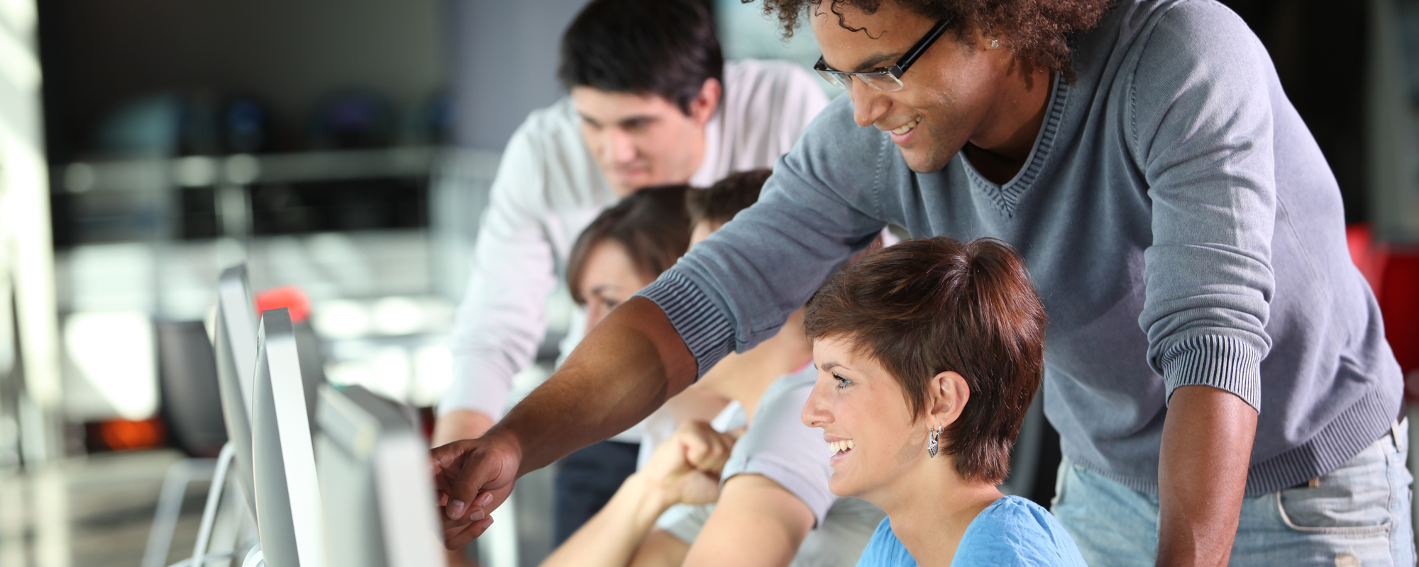 Male teaching a woman by pointing to something on her screen, both are laughing. There are others in the background.