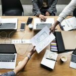 People sitting around a table with laptops in front of them while a paper is passed around