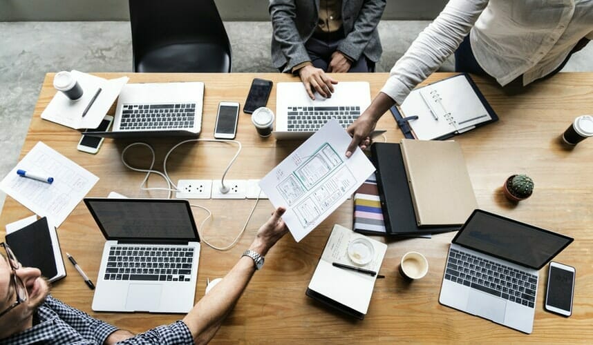 People sitting around a table with laptops in front of them while a paper is passed around