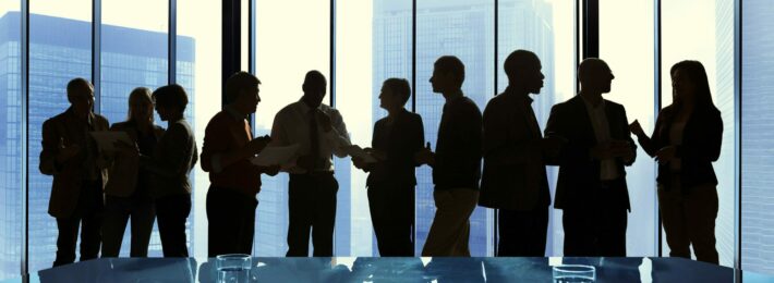 A silhouette of a line of co-workers standing and talking with each other in front of a high-rise window.