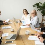 Serious aged businesswoman discussing corporate financial report with team talking to employees, senior mature female boss ceo leader analyzing work results with subordinates at company group meeting