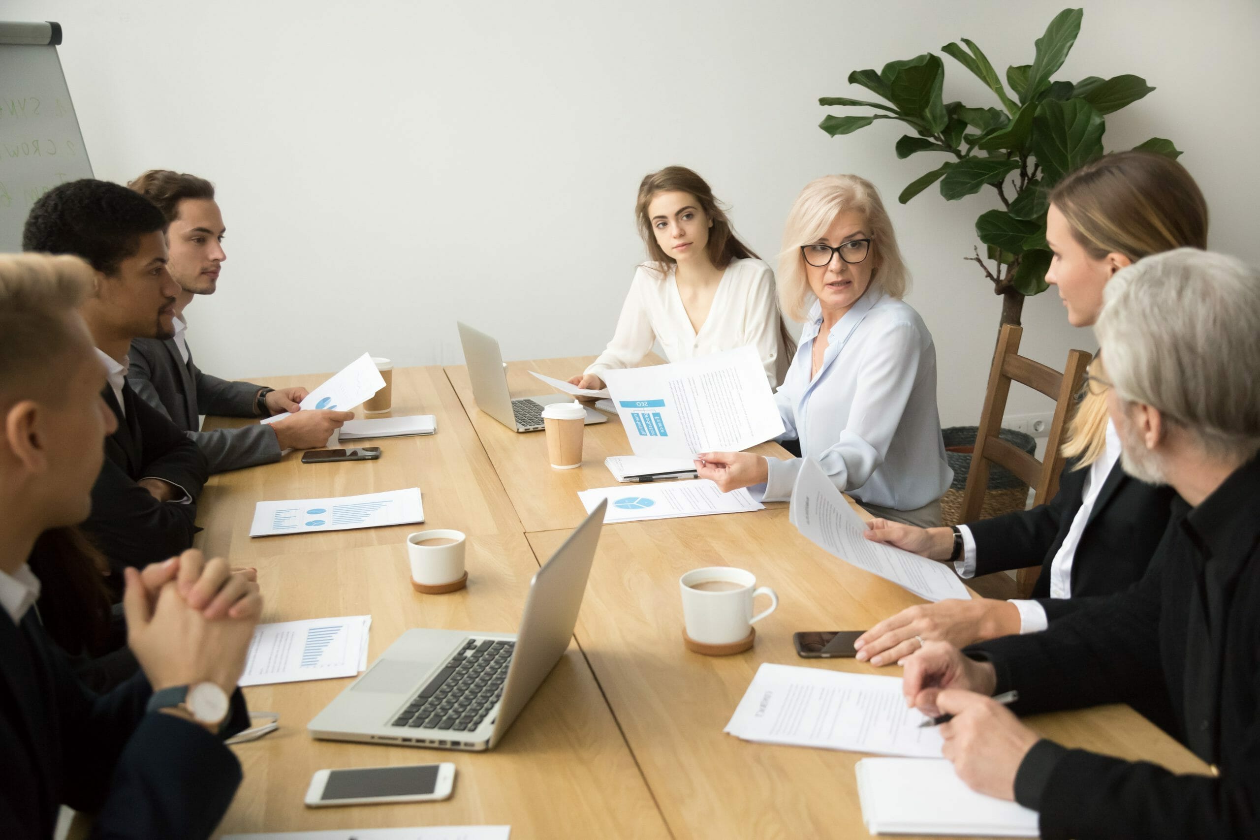 Serious aged businesswoman discussing corporate financial report with team talking to employees, senior mature female boss ceo leader analyzing work results with subordinates at company group meeting