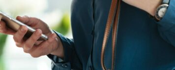 Side view portrait of a smiling young woman in eyeglasses using mobile phone while standing inside a cafe