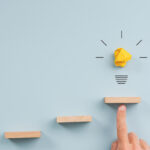 Hand supporting the top step in staircase of wooden pegs with a glowing light bulb above it.