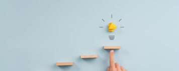 Hand supporting the top step in staircase of wooden pegs with a glowing light bulb above it.