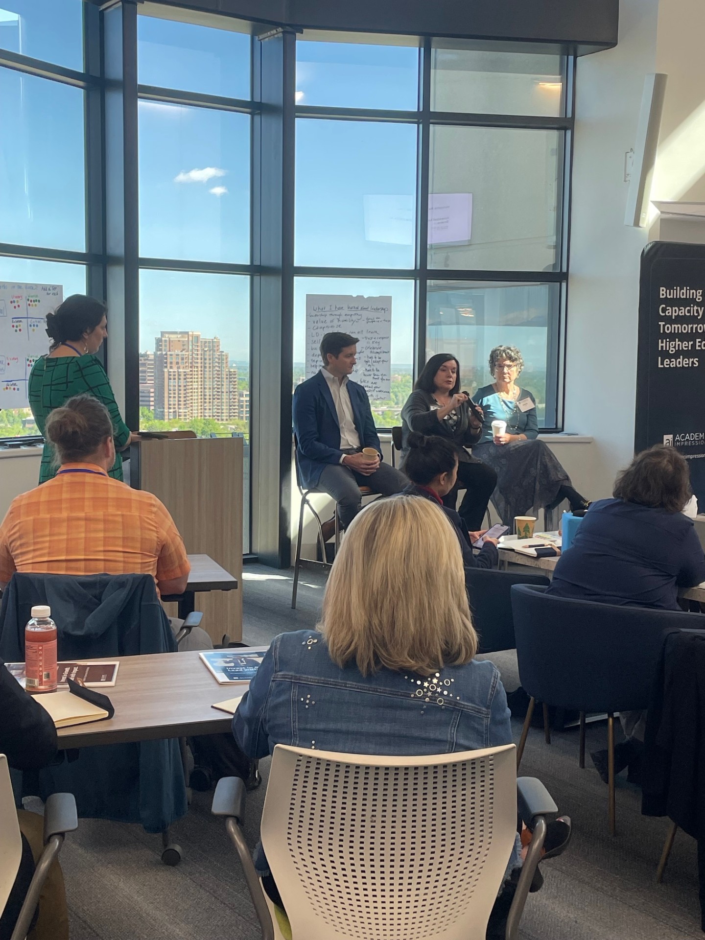 Image depicts an Academic Impressions workshop at their Denver office with 4 speakers up front and attendees sitting at tables listening to the speakers.