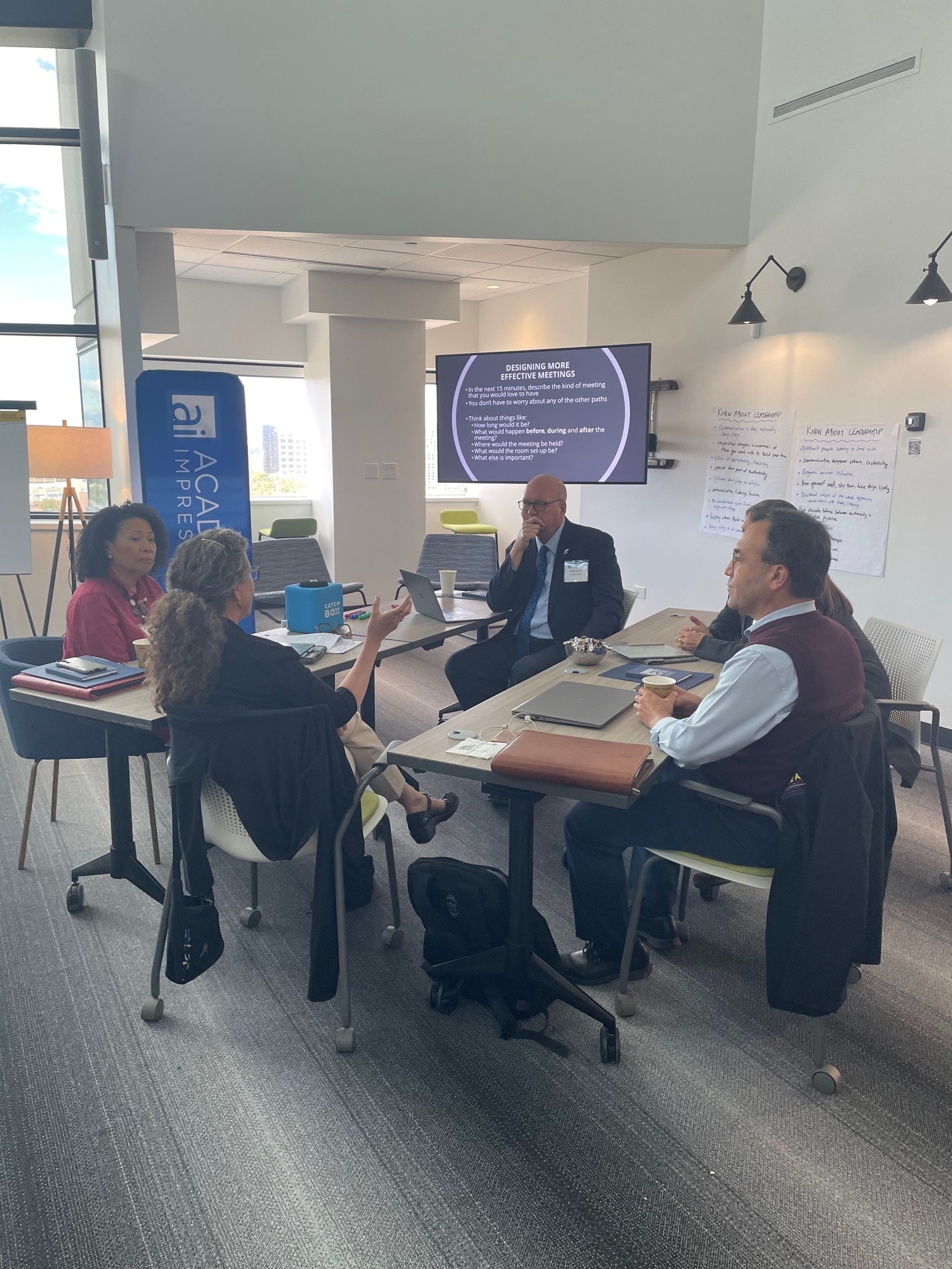 Four professionals seated around a table in a modern conference room, engaged in small group discussion during a leadership training workshop. A presentation slide titled 'Designing More Effective Meetings' is visible on a screen in the background.