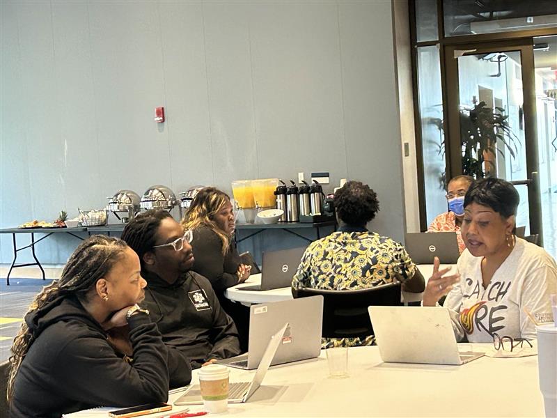  A group of six people seated around a conference table with laptops, engaged in discussion during a workshop or training session. A breakfast buffet is visible in the background.